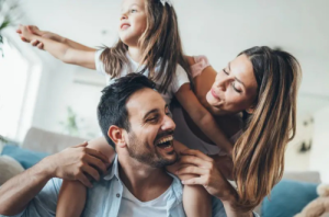 Smiling young girl sitting on father's shoulders, laughing and playing with mother standing behind.